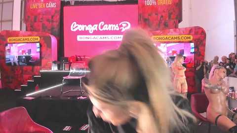 Media: Video of a woman with blonde hair in a ponytail, blurred, in front of a red and pink \"CamSoda\" stage set with two scantily-clad women, one holding a bottle, and another holding a microphone.