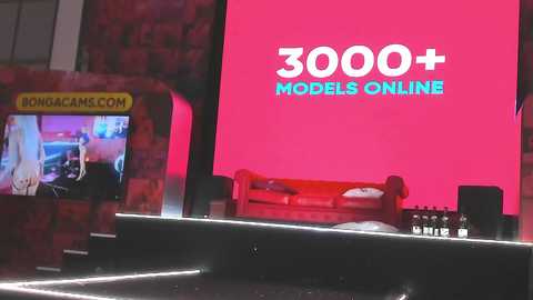 Media: Video of a bright pink screen displaying \"3000+ MODELS ONLINE\" text in white and blue, set against a dimly lit brick wall. A small TV shows a video, and a red sofa is visible in the background.