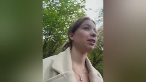 A video of a young Caucasian woman with light brown hair, wearing a beige coat, standing outdoors among lush green trees. Her expression is contemplative.