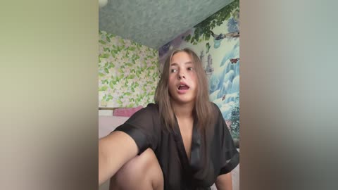 A young woman with light skin and long brown hair, wearing a sheer black top, sits on a bed with floral wallpaper, taking a selfie.