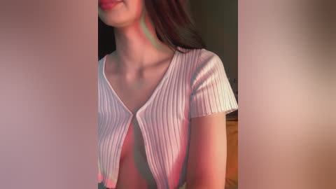 Video of a young woman with light skin, dark hair, wearing a white ribbed V-neck shirt. The background is blurred, with a warm, ambient light.