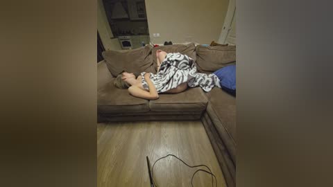 Video of a man and woman in a cozy living room, lying on a brown sectional sofa, wearing matching black and white patterned blankets, surrounded by wooden flooring and home decor.