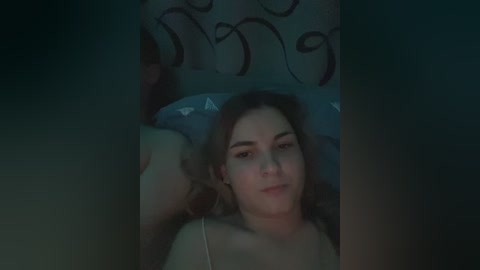 Video of a young woman with light skin and brown hair lying on a bed, wearing a light-colored top. Background features a bed with a dark, patterned headboard and a blue pillow. The lighting is dim, creating a moody atmosphere.