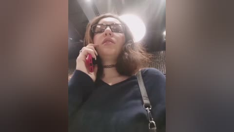 A video shows a woman with shoulder-length brown hair, glasses, and a black choker, speaking on a red phone while wearing a dark navy sweater. The blurred background suggests a car interior.