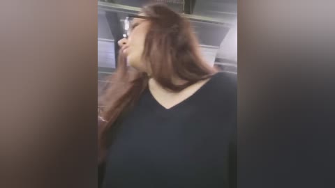 Video of a young Asian woman with long, straight brown hair, wearing a black V-neck sweater, standing in a dimly lit, blurry bathroom with white tiles and a glass shower door.