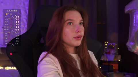 Video of a young, fair-skinned woman with long, straight brown hair, sitting in a dark gaming chair with a purple background. She wears a white top, and the room features a cozy, dimly lit ambiance with fairy lights and a calendar display.
