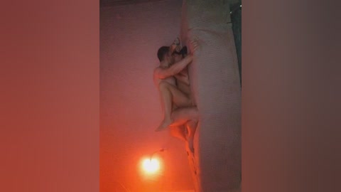 A video of a nude man and woman, both with light skin, embracing passionately on a white couch in a dimly lit, reddish-orange room.