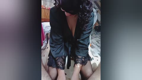 A video shows a person with dark curly hair, wearing a black satin robe with lace trim, kneeling on a gray carpeted floor in a dimly lit bedroom. The robe is open, revealing a black bra underneath.