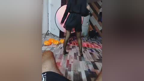 Video of a woman in black dress, stockings, and high heels dancing on a wooden floor with scattered orange fabric, in a room with a large light stand and wooden stairs.