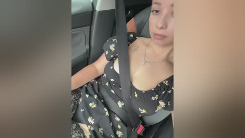 Video of a young woman with medium skin tone, wearing a black floral dress, reclined in a car seat with a seatbelt across her chest. She has a neutral expression and a small tattoo on her collarbone.