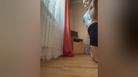 Video of a slim woman with fair skin, wearing a white sports bra and black shorts, kneeling on a wooden floor in a modest room with red curtains, lace window coverings, and a wooden shelf.
