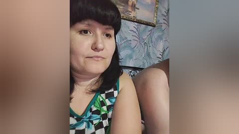 Video of a chubby, light-skinned woman with black hair, wearing a colorful patterned tank top, standing in a room with floral wallpaper and a framed painting in the background.