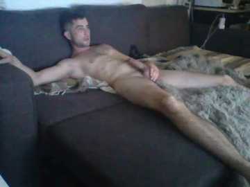 Video of a nude man with a circumcised penis, lying on a dark couch, covered in a gray blanket. Background shows a cluttered room with a white wall and a cluttered counter.