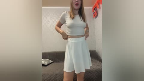 A video of a blonde woman with fair skin and medium build, wearing a white cropped top and high-waisted skirt, standing confidently in a beige room with a gray couch.