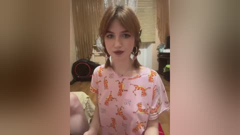 Media: Video of a young woman with light skin, brown hair, and freckles, wearing a pink t-shirt with cartoon animals, sitting in a cluttered bedroom with wooden blinds, a fan, and a bed visible.