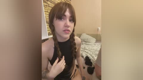 A video of a young woman with fair skin and long brown hair styled in braids, wearing a black top and black high-waisted shorts, sitting on a bed with a brick-patterned wall behind her.