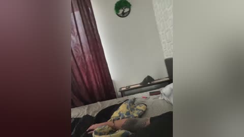 Video of a dimly lit, cluttered bedroom with a bed, a chair, and a green wall clock. The room has a dark curtain and a white textured wall.