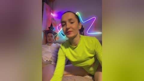 Video of a smiling, light-skinned woman in a neon green long-sleeve top, kneeling on a carpeted floor. Neon lights and a colorful backdrop with a cartoon character are visible in the background.