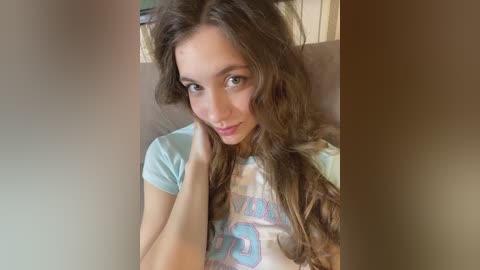 Video of a young Caucasian woman with long, wavy brown hair, light skin, and green eyes, wearing a pastel blue shirt with cartoon graphics. She has a soft, natural expression and is seated indoors, likely in a bedroom.
