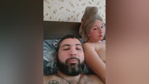 Video of a man and child on a bed, man with facial hair, child wrapped in a towel, floral wallpaper, casual setting.
