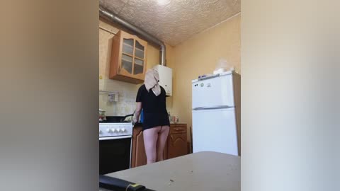 A video of a woman in a black top and white towel, cooking in a small, beige-walled kitchen with wooden cabinets and a white refrigerator.