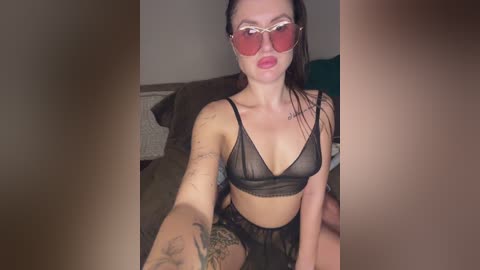 Video of a fair-skinned woman with dark hair, wearing red sunglasses and a sheer black bra, sitting on a brown couch.