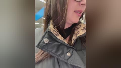 Media: Video of a woman with light skin, long brown hair, and glasses, wearing a black turtleneck under a grey puffy jacket with a beige lining. The background is blurry, suggesting an indoor setting.