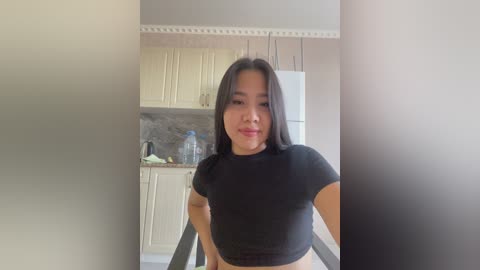 Video of a young Asian woman with long black hair, medium skin tone, wearing a black crop top, standing in a modern kitchen with white cabinets and a gray backsplash.