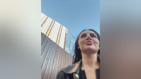 Media: Video of a young woman with long dark hair, wearing a black leather jacket, standing in front of a tall, modern white building with a clear blue sky.