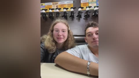 Media: Video of two young people, a girl with long curly hair in a leather jacket, and a boy with short brown hair in a white shirt, seated at a table in a room with a shelf displaying multiple toy figures.
