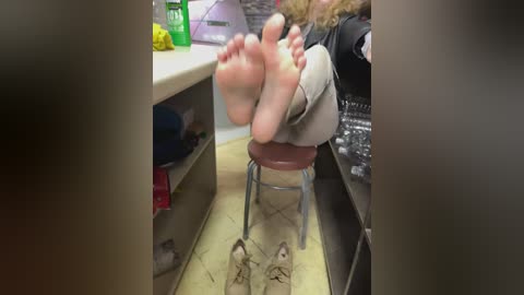 Media: Video of a woman with curly hair, barefoot, sitting on a red chair in a kitchen, legs crossed, feet facing the camera. Background includes kitchen counters, yellow flowers, and a green bottle.