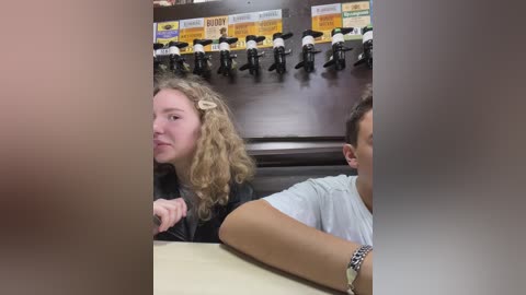 Media: Video of two young people, a blonde woman with curly hair and a man with short hair, sitting at a table in a dimly lit bar. They are facing away from the camera.