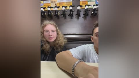 Media: A video shows two people in a bar; a blonde woman with curly hair in a black jacket, and a man in a white t-shirt, both sitting at a wooden bar. Behind them, beer taps are mounted on a dark wood wall.