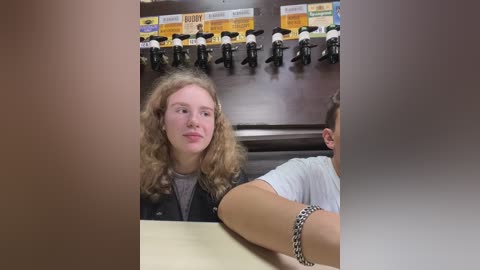 Media: Video of a young woman with long, curly blonde hair in a casual setting, wearing a gray shirt. Background shows a row of microphones and yellow signs.
