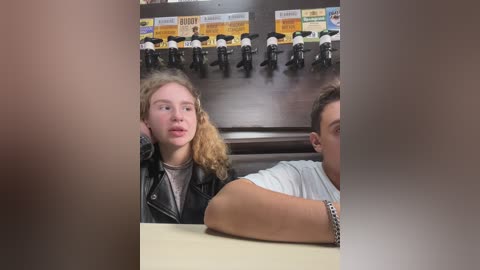 Media: Video of two young people, a blonde girl with curly hair in a black leather jacket and a boy with short hair in a white t-shirt, sitting in a fast food restaurant with a wall of motorcycle posters.