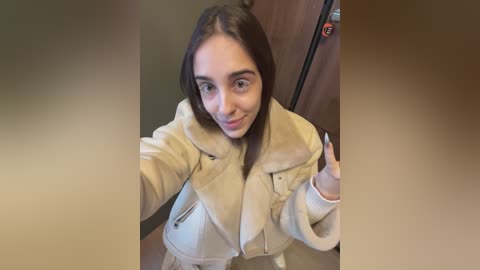 Media: Video of a young woman with light skin, straight brown hair, and a light beige jacket, smiling while taking a selfie in a wooden room.