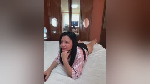 A video of a woman lying on a bed in a hotel room. She has long black hair, fair skin, and is wearing a pink and white striped shirt and black shorts. The room features wooden wardrobes and a TV in the background.