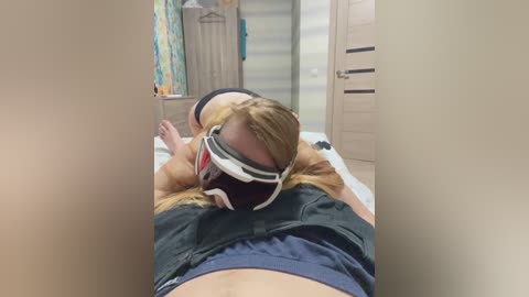 Video of a blonde woman lying on a bed in a bedroom, wearing a black and white headband and a dark shirt, with a blurred background showing a door and a dresser.