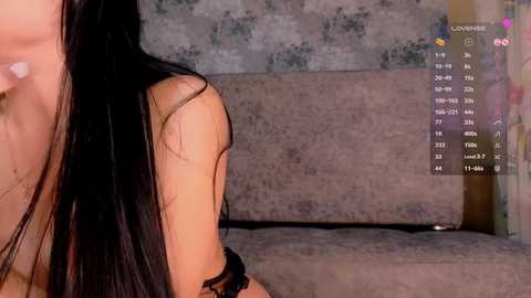 Video of a woman with long black hair, wearing a black top, sitting on a beige couch against a textured grey wall. Her back is visible, with a \"Lovense\" logo overlay displaying metrics.