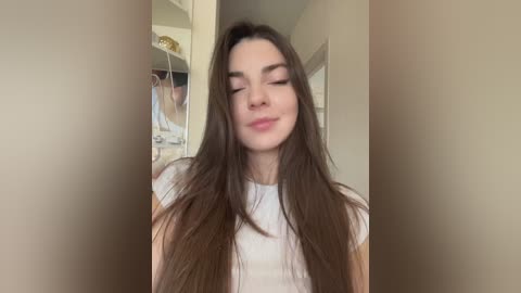 Media: Video of a young Caucasian woman with long, straight brown hair, wearing a white T-shirt, smiling softly, standing in a dimly lit, partially open closet with hanging clothes.
