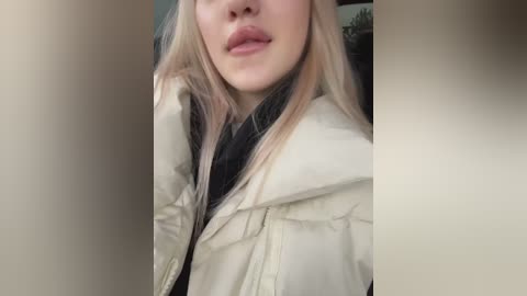 Media: A video of a young woman with light skin, long blonde hair, and a slightly open mouth, wearing a beige jacket, taken from a close-up angle, with a blurry background.