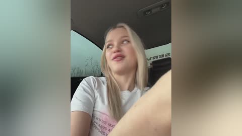 Media: Video of a young, blonde woman with fair skin, wearing a white t-shirt with text, sitting in a car. She has a contemplative expression, looking out the window.