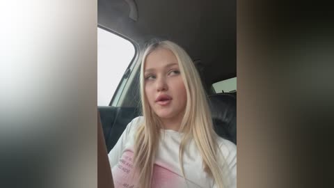 Media: Video of a blonde woman with fair skin and a soft expression, wearing a white top, sitting in a car with a blurred window and black leather seat in the background.