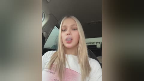 Media: Video of a blonde teenage girl with long hair, wearing a white t-shirt, sticking her tongue out, taken from the front seat of a car.