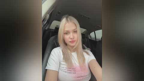 Media: Video of a blonde young woman with light skin, wearing a white t-shirt with a text print, sitting in a car, making a pouty face.