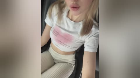 Media: A video of a young woman with light skin, blonde hair, wearing a tight white crop top revealing her midriff, and high-waisted cream-colored pants, sitting in a car.