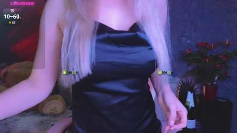 Video of a slender, fair-skinned woman with long, platinum blonde hair, wearing a black satin dress, indoors with a dimly lit, cozy ambiance, featuring a red flower arrangement in the background.