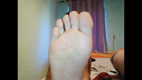 A video featuring a close-up of a pale, soiled foot, with the sole facing the camera. The background shows a bedroom with a purple curtain and a bed with white sheets.