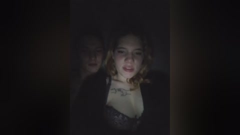 A dimly lit video shows a young woman with curly hair, wearing a black lace bra, partially obscured by a shadowy figure behind her. The background is dark and indistinct.