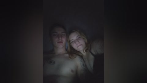 A dimly lit video featuring two young women, one topless with small breasts and tattoos, embracing in a dark, shadowy room.